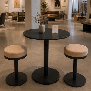 May include: A modern black round table with two matching stools. The stools have tan upholstered seats and black bases. A small vase with dried flowers sits on the table. The scene is set in a well-lit showroom.