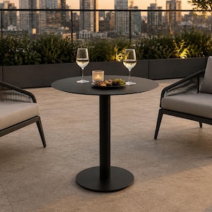 Puede incluir: Juego de patio al aire libre con una mesa redonda negra, dos sillones con cojines grises y dos copas de vino. Una pequeña vela y un plato de comida están sobre la mesa. El fondo muestra un paisaje urbano al atardecer.