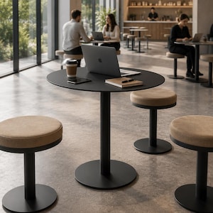 Può includere: Un moderno ambiente da caffè con tavoli rotondi neri e sgabelli abbinati. Gli sgabelli hanno sedute imbottite beige. Un laptop, un taccuino e una tazza di caffè sono su un tavolo. Le persone sono sedute ai tavoli sullo sfondo, che lavorano sui laptop.