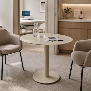 Op de afbeelding: Een moderne eethoek met een ronde, crèmekleurige tafel met een enkele voet, geflankeerd door twee gestoffeerde fauteuils met zwarte metalen poten. Een glazen karaf en glazen staan op de tafel, wat wijst op een eet- of wachtruimte.