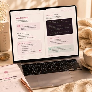 May include: A laptop screen displaying a coding tutorial titled Mood Checker with text about building a programme, alongside a spiral-bound notebook titled The Coding Journal, a cup of coffee, and a vase of dried flowers on a desk surface.