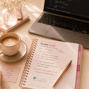 May include: A spiral-bound weekly planner open to a page titled Weekly Goals, featuring sections for top goals, learning objectives, and a task list. A rose gold pen rests on the page, which includes text about Python functions and a Tip Splitter project.