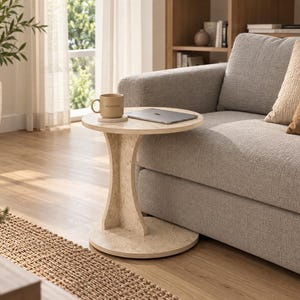 May include: A round side table with a light beige speckled finish, featuring a circular top and a curved central support column that connects to a circular base. The table is positioned next to a grey sofa and holds a mug and a laptop.