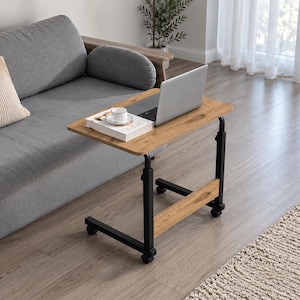 Puede incluir: Una mesa auxiliar móvil con un tablero rectangular de madera clara y una estructura de metal negro. La mesa cuenta con cuatro ruedas pequeñas en la base, una estructura ajustable en altura y una barra de soporte horizontal de madera.