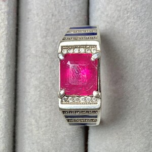 May include: A silver ring featuring a large, rectangular, vibrant pink gemstone held by four prongs. The band is accented with rows of small, clear, round stones and horizontal dark blue enamel stripes along the sides of the metal setting.