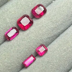 May include: A collection of five loose, faceted gemstones in vibrant shades of pink and red. The stones feature various cuts, including rectangular, square, and cushion shapes, displaying clear facets and reflective surfaces arranged on a gray tray.