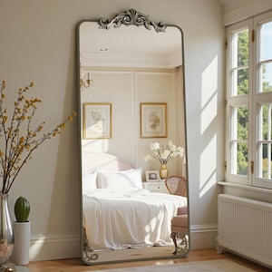 May include: A tall, rectangular mirror with an ornate, silver-colored decorative top and bottom. The mirror has a thin, gray frame. The reflection shows a bedroom with a bed, artwork, and a window. The room is painted in neutral colors.