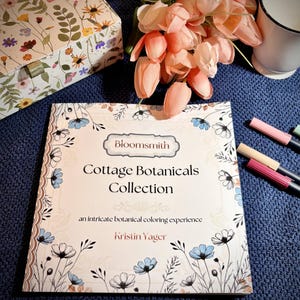 May include: A floral-themed coloring book titled "Cottage Botanicals Collection" by Kristin Yager, with the "Bloomsmith" logo. The book is surrounded by pink and beige markers, a floral box, and a bouquet of pink tulips on a blue surface.