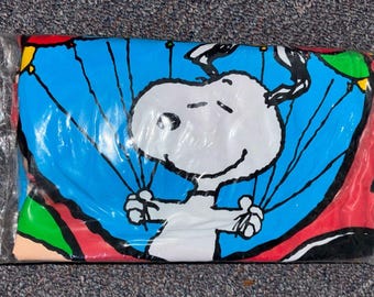 May include: A folded t-shirt sealed in a clear plastic bag featuring a graphic of the cartoon character Snoopy. The design shows a white Snoopy against a bright blue circular background, holding onto black strings attached to colourful balloons.
