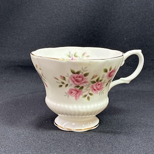 May include: A white porcelain teacup featuring a delicate pink rose floral pattern with green leaves. The cup has a scalloped rim, a ribbed base, and a gold-trimmed handle and foot. The interior also displays a small pink rose motif.
