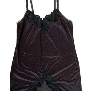 May include: A black slip dress featuring a smooth, satin-like fabric with adjustable shoulder straps. The garment is accented with intricate black floral lace embroidery along the V-neckline and a matching lace detail at the center hemline.
