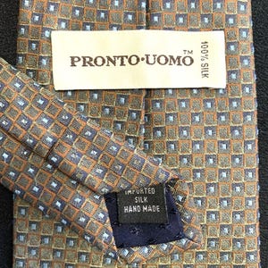 May include: A necktie featuring a repeating geometric pattern of orange and blue squares with small light blue dots. A white label reads PRONTO UOMO 100% SILK, and a black label reads IMPORTED SILK HAND MADE. The tie is constructed from silk fabric.