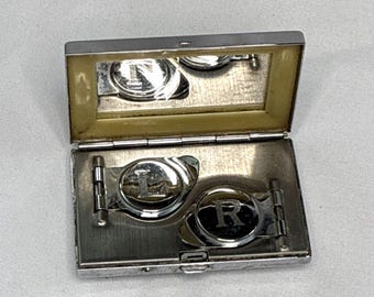 May include: A rectangular silver-toned metal case with a hinged lid and an interior mirror. Inside the case are two circular silver-toned metal clips, each engraved with a letter, one featuring an L and the other featuring an R, secured by hinges.
