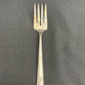 Könnte beinhalten: Eine silberne Gabel mit vier Zinken und einem dekorativen Griff. Die Gabel hat ein verziertes Design am Griff und ist vor einem dunklen Hintergrund platziert. Die Zinken sind leicht gebogen.