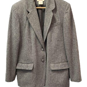 May include: A grey wool-blend blazer featuring a notched lapel, two dark buttons for closure, and two front flap pockets. The garment is lined with black fabric and includes a brand label at the inner neckline, all displayed on a wooden hanger.
