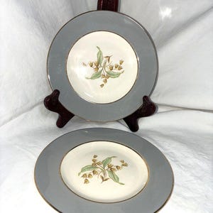 May include: Two round ceramic plates featuring a cream-coloured centre with a botanical illustration of green leaves and small brown bell-shaped flowers. The plates have a wide grey border and a thin gold rim around the edge of the centre and outer rim.