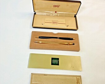 May include: A Cross pen set featuring one black pen and one gold-tone pen, presented in a brown hinged case with a beige interior. The set includes a guarantee and operating instructions card, a gold-coloured information card with the Cross logo.