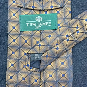 May include: A gold and brown necktie featuring a dark blue grid pattern with small blue squares at each intersection. A green label reads Tom James, and a smaller black tag indicates the material is all silk. The tie has a pointed end.
