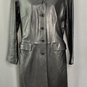 May include: A black leather coat with a notched collar, long sleeves, and a button-down front. The coat features two front pockets and a tailored fit. The coat is displayed on a mannequin against a neutral background.