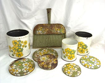 May include: A collection of vintage metal kitchenware including two cylindrical canisters, a dustpan, and four circular lids. Each item features a white or green base with a repeating yellow and green floral pattern and visible signs of rust.