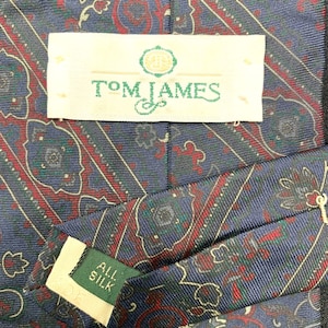 May include: A blue necktie featuring a repeating paisley pattern in red, green, and gold. A white label stitched to the back displays the text Tom James with a logo. A smaller green tag tucked into the fold of the tie reads All Silk in white text.