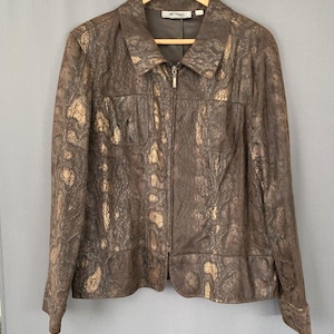 Puede incluir: Una chaqueta de manga larga con un patrón de piel de serpiente en color marrón oscuro y dorado metálico. La chaqueta incluye una cremallera frontal completa, un cuello en punta y dos bolsillos en el pecho. La prenda cuelga de una percha de madera.
