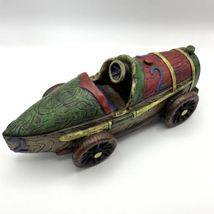 May include: A toy race car featuring a wood-grain texture in shades of brown, green, and red. The vehicle has four dark brown wheels, a small steering wheel inside the cockpit, and the number 2 painted in blue on the front and top sections.