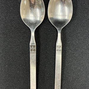 May include: Two silver-toned metal spoons with oval bowls and flat handles. Each handle features a decorative band with a repeating four-petal floral pattern located just below the neck of the spoon, leading down to a plain, straight-edged stem.