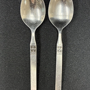 May include: Two silver-toned metal spoons with a polished oval bowl and a flat, straight handle. Each handle features a decorative band near the neck containing a repeating pattern of four-petaled flower motifs embossed into the metal surface.