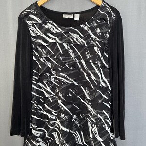 May include: A long-sleeved black top featuring a scoop neckline and a central panel with horizontal, layered ruffles. The ruffled section displays a black, white, and gray abstract brushstroke pattern. The garment is displayed on a wooden hanger.