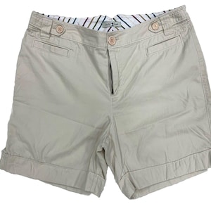 May include: Beige shorts featuring a button-front closure, two side pockets, and two decorative buttons at the waistband. The interior waistband is lined with a white fabric featuring thin, multicolored vertical stripes. The Duck Head brand label is visible.