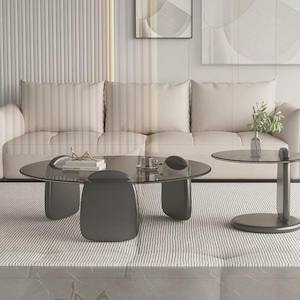 Puede incluir: Una mesa de centro redonda grande con tapa de cristal oscuro y tres patas redondeadas de color gris oscuro, junto a una mesa auxiliar a juego más pequeña con base de pedestal, ambas con acabado liso y diseño estructural moderno.