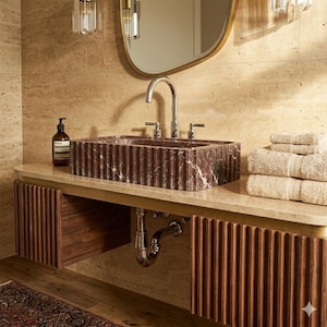 May include: A rectangular, fluted dark red marble sink sits on a light beige stone countertop. Below, a floating wooden vanity features vertical slats. A chrome faucet, a brown soap bottle, and a stack of beige towels are visible nearby.