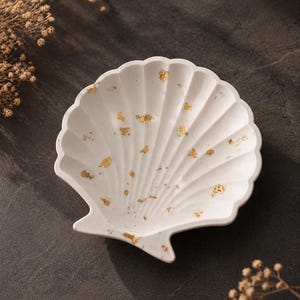May include: A white shell-shaped trinket dish featuring a scalloped edge and a textured surface embedded with small, scattered gold leaf flakes. The dish has a concave center and a defined hinge area at the base, showcasing a clean, bright finish.