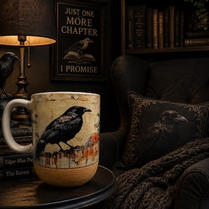 May include: A ceramic mug with a white handle and a cork base, featuring a detailed illustration of a black raven perched on a textured, aged-paper background with orange accents. The mug displays a dark, artistic aesthetic with visible paint drips.