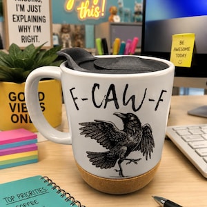 May include: A white ceramic mug with a black lid and a cork base. The mug features a black ink illustration of a crow in flight and the text F-CAW-F printed in a bold, hand-drawn style font on the front surface of the white ceramic body.