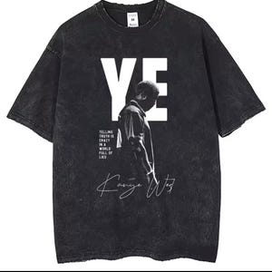 Op de afbeelding: Een zwart acid-wash t-shirt met een grote witte "YE"-afbeelding, een zwart-wit silhouet van een persoon, de tekst "TELLING TRUTH IS CRAZY IN A WORLD FULL OF LIES" en een witte handgeschreven handtekening "Kanye West".