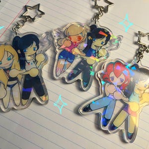 Peut inclure: Trois porte-clés en acrylique transparent représentant des personnages de dessin animé avec des fermoirs métalliques en forme d'étoile. Chaque porte-clé montre deux personnages aux tenues et cheveux variés, avec une finition holographique brillante.