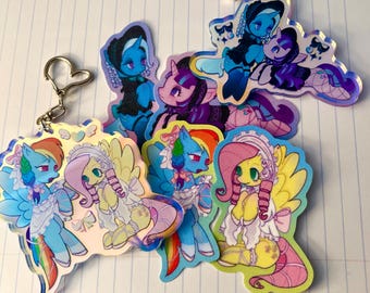 May include: A collection of acrylic charms featuring stylized pony characters in various outfits. One charm includes a heart-shaped metal keyring. The designs showcase colorful ponies with detailed manes, lace-trimmed clothing, and accessories in a vibrant art style.