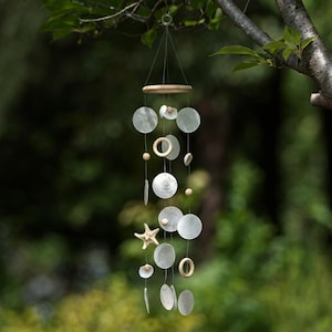 May include: A hanging mobile featuring circular white shell discs, small wooden rings, and a central starfish. The components are suspended by thin wires from a circular wooden frame, creating a layered arrangement of natural materials and textures.