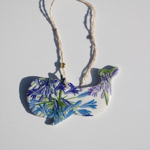 May include: A whale-shaped ornament featuring a white base with a vibrant blue and green floral pattern. The ornament is suspended by a thin, light-coloured twine string that includes a small, round, metallic gold bead for decorative detail.