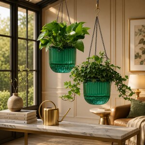 May include: Two emerald green ribbed glass hanging planters suspended by dark metal chains, each containing a lush green plant. A gold metal watering can sits on a white marble table next to a stack of three books and a small beige ceramic vase.