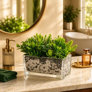 May include: A rectangular clear glass planter filled with a mixture of small white and gray pebbles, topped with vibrant green artificial foliage. The planter sits on a marble surface next to a ribbed gold soap dispenser and a glass perfume bottle.