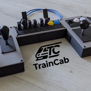 Könnte beinhalten: Eine modulare Zugsimulator-Steuerkonsole mit grauem und schwarzem Gehäuse, ausgestattet mit mehreren Joysticks, Hebeln und Tasten. Das Gerät zeigt den Text TrainCab sowie den Satz YOUR controller for nearly every train simulator.