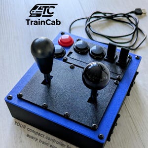 Könnte beinhalten: Ein blau-schwarzer Zugsimulator-Controller mit zwei schwarzen Joysticks, einem roten Knopf, drei schwarzen Knöpfen und zwei schwarzen Kippschaltern. Das Gerät enthält ein USB-Kabel und zeigt den Text TrainCab sowie YOUR compact controller for nearly every train simulator.