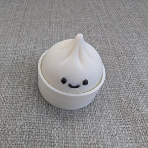 Op de afbeelding: Een witte 3D-geprinte container in de vorm van een dumpling met een lachend gezicht. De container heeft een deksel dat lijkt op de bovenkant van een dumpling. De basis van de container heeft de vorm van een ronde kom. De dumpling heeft een getextureerd oppervlak.