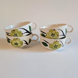 May include: Four cream-colored ceramic cups arranged in two stacks of two. Each cup features a hand-painted design of a large, light green flower with dark green leaves and small red accents. The cups have a rounded shape and a side handle.