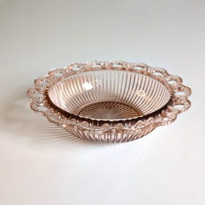 May include: A pink glass bowl with a scalloped edge and vertical ribbed detailing. The bowl has a circular shape and a delicate, vintage aesthetic. The bowl is likely used for serving or display.
