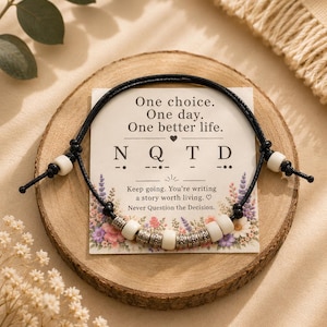 Peut inclure: Un bracelet en cordon noir avec des perles blanches et argentées présenté sur une carte carrée portant le texte : One choice. One day. One better life. N Q T D. Keep going. You're writing a story worth living. Never Question the Decision.
