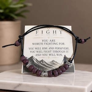 Könnte beinhalten: Ein Armband mit dunkelvioletten Perlen und silberfarbenen Akzenten wird vor einer Karte mit dem Wort "FIGHT" und inspirierendem Text präsentiert. Das Armband liegt auf einer weißen Marmoroberfläche.